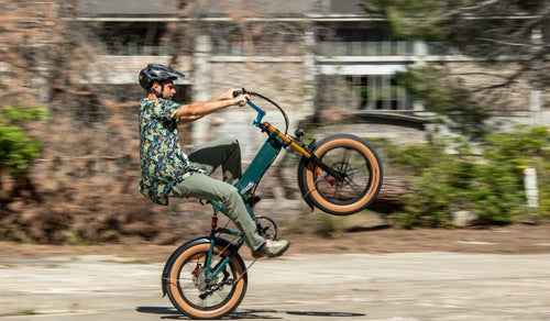 Un ciclista esegue un’impennata su una fat e-bike Fiido M1 Pro, mettendo in risalto il telaio pieghevole compatto, gli pneumatici larghi e la sospensione anteriore su una strada urbana.