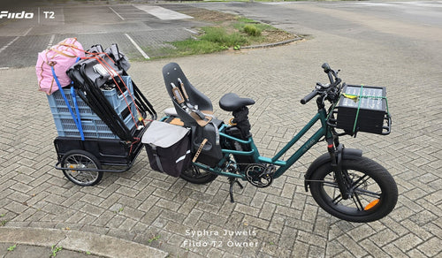 Una cargo e-bike Fiido T2 è mostrata completamente carica, con un seggiolino per bambini, borse laterali, un box di carico anteriore e un rimorchio posteriore carico di bagagli.